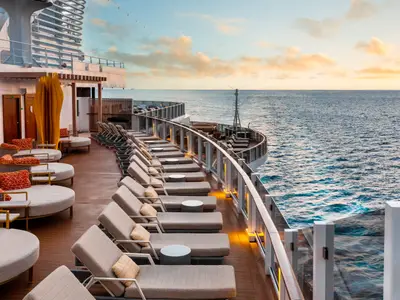 Ein weitläufiges Sonnendeck an Bord der Norwegian Prima bei Sonnenuntergang. Reihen eleganter Liegestühle und runder Daybeds bieten Meerblick unter einem warmen Himmel.
