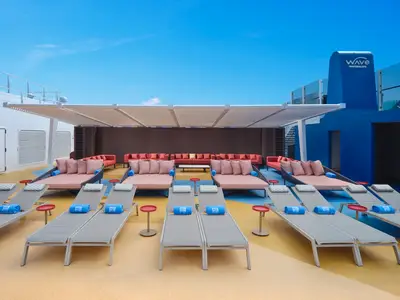 Sonnendeck an Bord der Norwegian Prima mit bequemen Liegen, gepolsterten Tagesbetten und der blauen Wave Wasserrutsche im Hintergrund unter strahlend blauem Himmel. Blaue Handtücher mit NCL-Logo liegen auf den Liegen bereit.