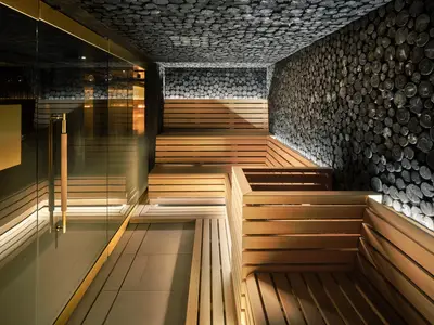 Innenansicht einer luxuriösen Holzkohle-Sauna an Bord der Norwegian Prima. Dunkle Wände aus Holzscheiben und helle Holzpritschen in mehreren Ebenen, beleuchtet von warmem Licht. Eine Glastür mit goldenem Griff ist links sichtbar.