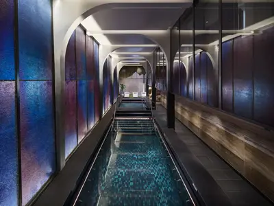 Der lange Spa-Pool-Bereich an Bord der Norwegian Prima. Die Wände sind mit großen, farbigen Paneelen in Blau und Violett gestaltet. Zwei moderne Pools und bequeme Liegen sind zu sehen.