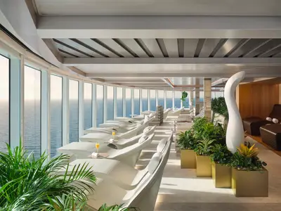 Die helle Spa Thermal Lounge an Bord der Norwegian Prima mit modernen weissen Liegen und grossen Panoramafenstern zum Meer. Grüne Pflanzen und eine weisse Skulptur schmücken den Raum.