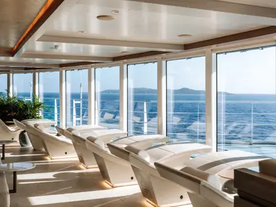 Elegante Thermal Lounge der Norwegian Prima. Moderne, helle Liegen vor Panoramafenstern mit Blick auf das blaue Meer und grüne Inseln.