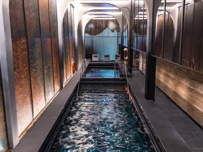 Ein eleganter Thermalpool im Spa-Bereich der Norwegian Prima. Der dunkle Pool mit blauer Unterwasserbeleuchtung ist von kunstvollen Wänden und Decken umgeben. Zwei Rettungsringe sind an den Wänden zu sehen. Ohne Personen.