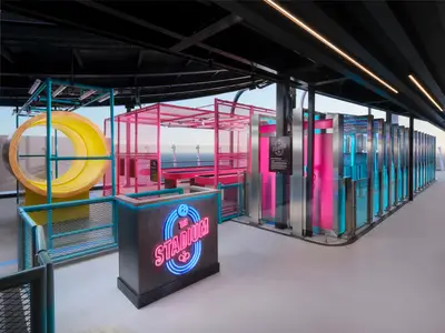 Detailansicht des "The Stadium" Sport- und Aktivitätsbereichs an Bord der Norwegian Prima mit leuchtenden Neon-Farben, einer gelben Röhre, rosafarbenen Käfigen und blauen Lichtinstallationen. Im Hintergrund ist das Meer zu sehen.