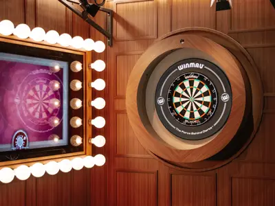 Ein moderner Dartbereich namens "The Bulls Eye" an Bord der Norwegian Prima. Zu sehen sind ein beleuchtetes, elektronisches Dartboard in einer Holzumrandung und ein großer Bildschirm daneben, der ebenfalls eine digitale Dartscheibe anzeigt. Beide Elemente sind von zahlreichen hellen Glühbirnen eingerahmt und die Wand ist mit Holzpaneelen verkleidet.