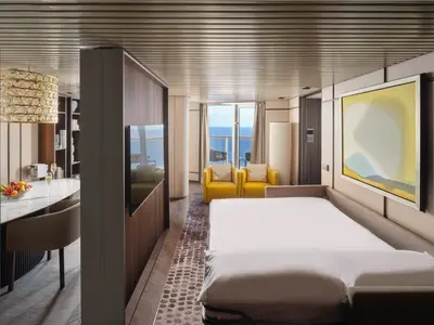 Modern suite on the Norwegian Prima with a king-size bed, two yellow armchairs, dining area, and a balcony with ocean views.