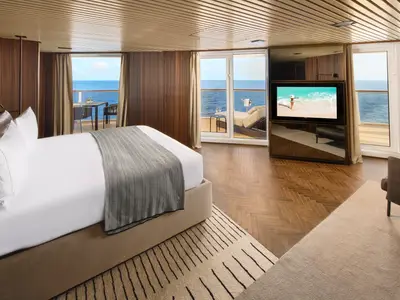 Elegante Suite an Bord der Norwegian Prima mit großem Doppelbett, Holzboden und Balkon mit Meerblick. Ein Fernseher und eine gemütliche Sitzecke sind ebenfalls sichtbar.