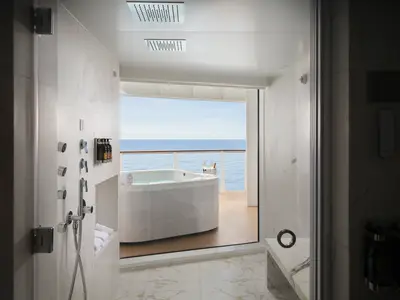 Ein luxuriöses Badezimmer an Bord der Norwegian Prima mit einer modernen Dusche aus Marmor. Durch die Glasfront blickt man auf einen Balkon mit Whirlpool, Sekt und weitem Meerblick.