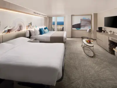 Geräumige Familiensuite an Bord der Norwegian Prima mit zwei großen Betten, Wohnbereich, Balkon mit Meerblick. Moderne Kunst, Obst und Sekt sind ebenfalls zu sehen.