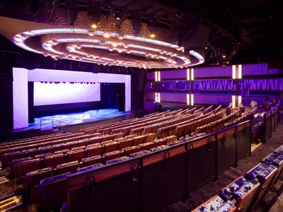 Weitwinkelaufnahme des stilvollen Theaters an Bord der Norwegian Prima mit leuchtender Bühne, leeren Sitzreihen und futuristischer lila Beleuchtung.