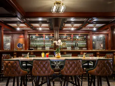 Eine Innenansicht der eleganten Sugarcane Mojito Bar an Bord der Norwegian Sky. Man sieht eine lange Bar mit vielen Spirituosenregalen und davor Sitzgelegenheiten mit braunen Ledersesseln und einem Holztisch.
