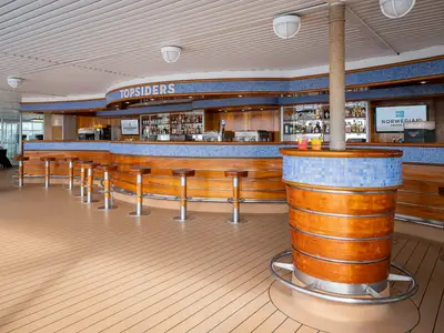 Die Topsiders Bar an Bord der Norwegian Sky, mit Holztheke, blauen Mosaikfliesen und Barhockern. Flaschenregale und NCL-Bildschirm im Hintergrund. Deck in Holzoptik.