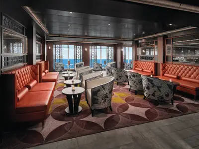 Panoramablick auf die stilvolle Pinnacle Lounge an Bord der Norwegian Sky. Orangefarbene gepolsterte Sofas und Sessel mit Blattmuster vor großen Fenstern mit Meerblick schaffen eine gemütliche Atmosphäre.