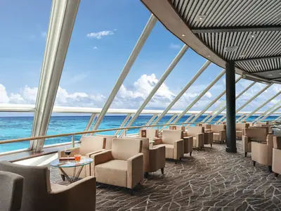 Spinnaker Lounge an Bord der Norwegian Sky. Helle Fensterfront mit Blick auf das türkise Meer. Beige Sessel, Glastische und Drinks.