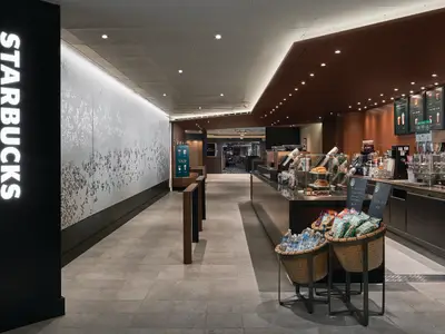 Starbucks-Café an Bord der Norwegian Sky. Ein langer Tresen mit Kaffeemaschinen, Gebäckvitrinen und Getränkeanzeigen. Der Gang zum Café ist mit zwei Körben voller Snacks und Wasserflaschen gesäumt. Eine elegante Wanddekoration ist links zu sehen, während im Hintergrund ein Casino erkennbar ist.