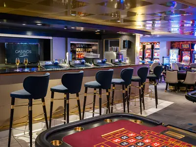 Das Sky Club Casino an Bord der Norwegian Sky. Eine elegante Bar mit blauen Hockern und zwei Champagnergläsern. Im Hintergrund leuchten Spielautomaten und Tische. Eine abendliche Unterhaltungsszene.
