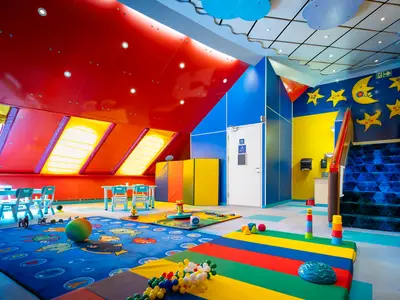 Bunter Kinderbereich "Guppies Room" an Bord der Norwegian Sky. Rote Wände, blaue Decke, gelbe Fenster. Spielmatten, Kindermöbel, Spielzeug. Sterne und Mond an Wand, rechts blaue Treppe.