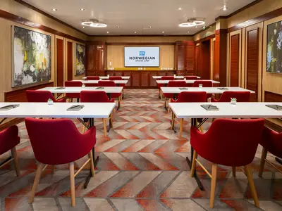 Ein modern eingerichteter Konferenzraum an Bord der Norwegian Sky mit weißen Tischen und roten Stühlen, ausgerichtet auf eine große Leinwand mit dem NCL-Logo. Auf den Tischen liegen Notizbücher.