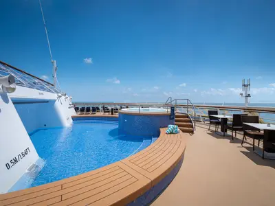 Poolbereich an Deck der Norwegian Sky. Blauer Pool und Whirlpool. Holzdeck mit Handtüchern und Sitzen. Weiter Ozean unter blauem Himmel im Hintergrund. Wassertiefenangaben sichtbar.