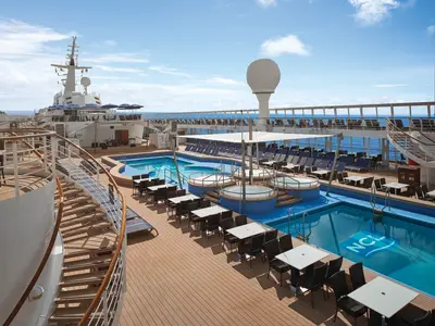 Weitwinkelansicht des sonnigen Pooldecks der Norwegian Sky mit zwei blauen Pools, mehreren Whirlpools und Reihen von Liegestühlen. Im Hintergrund ist das blaue Meer zu sehen.