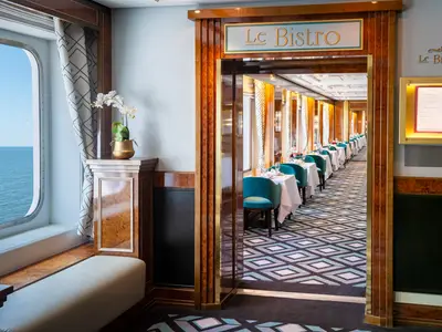 Das Le Bistro Restaurant an Bord der Norwegian Sky. Eine lange Reihe elegant gedeckter Tische mit tealfarbenen Stühlen erstreckt sich in einem Raum mit dunklem Holzdekor und einem Fenster mit Meerblick.