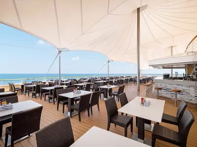 Einladender Aussenbereich des Restaurants The Great Outdoors an Bord der Norwegian Sky. Zahlreiche Tische, Stühle und eine Bar unter einem grossen Sonnensegel. Der blaue Ozean und klare Himmel im Hintergrund. Keine Personen.