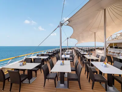 Blick auf das offene Deckrestaurant The Great Outdoors an Bord der Norwegian Sky. Zahlreiche Tische und Stühle stehen unter einem großen Sonnensegel, im Hintergrund ist das weite Meer zu sehen. Rechts eine Bar.