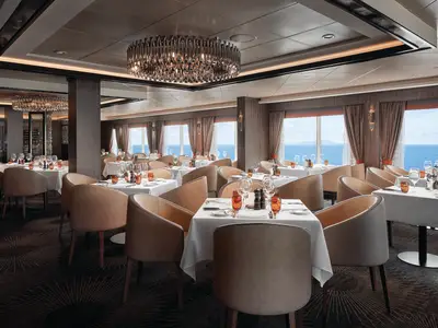 Blick in das moderne Cagney's Steakhouse an Bord der Norwegian Sky mit vielen hellbraunen Stühlen, weiß gedeckten Tischen und großen Fenstern zum Meer. Eine aufwendige Lichtinstallation hängt an der Decke.