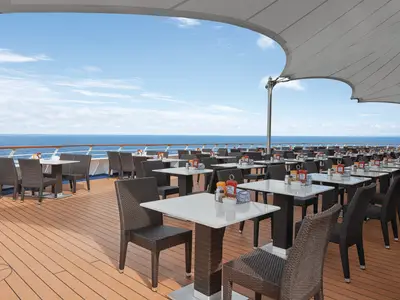 Der Aussenbereich eines Restaurants auf der Norwegian Sky mit zahlreichen Tischen und Stühlen und weitem Blick über das Meer unter blauem Himmel.