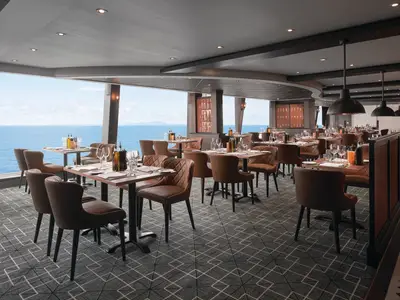 Ein elegantes italienisches Spezialitätenrestaurant an Bord der Norwegian Sky mit vielen Tischen, braunen Ledersesseln und einem weiten Meerblick durch Panoramafenster.