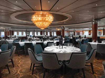 Hauptspeisesaal an Bord der Norwegian Sky. Mehrere Tische mit weißen Tischdecken und Stühlen. Ein großer goldener Kronleuchter hängt von der Decke. Durch Fenster ist das Meer zu sehen.