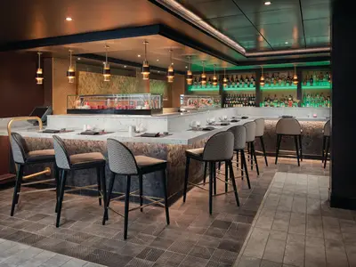 Eine elegant beleuchtete Sushi-Bar mit langer Theke und Barhockern, flankiert von einer Spirituosenbar, an Bord der Norwegian Sky. Moderne Einrichtung mit Holz- und Steinakzenten.