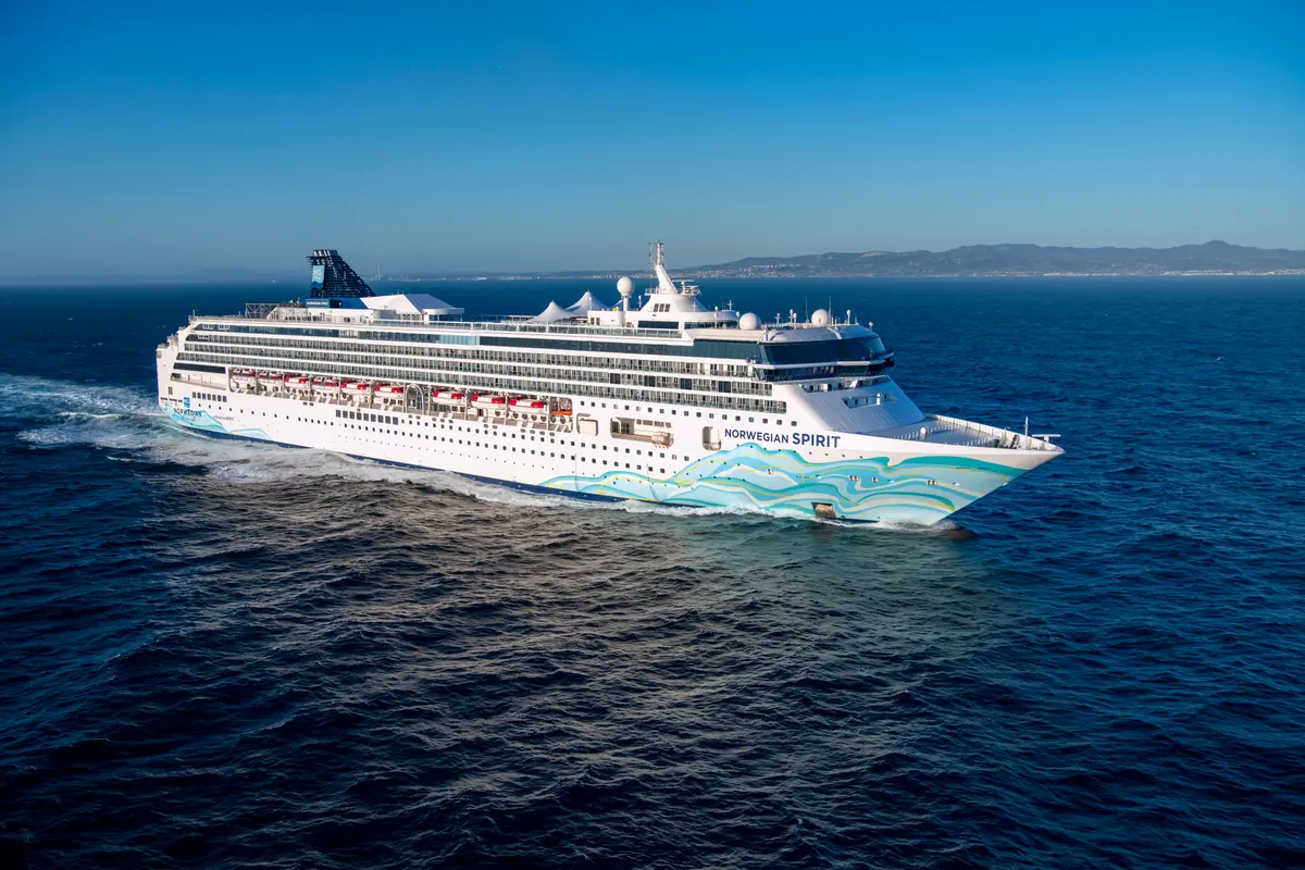 Die Norwegian Spirit fährt auf dem blauen Meer unter klarem Himmel. Das weiße Schiff mit den blauen und grünen Wellenmustern am Bug hinterlässt eine Schaumspur. Im Hintergrund sind entfernte Berge zu sehen.