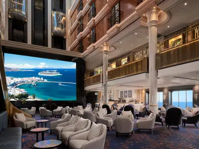Modernes Atrium der Norwegian Spirit mit grosser LED-Wand, die Mykonos zeigt. Komfortable Sessel und eine Café-Bar prägen den Lounge-Bereich. Aufzüge oben.
