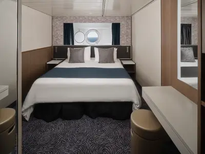 Helle Aussenkabine an Bord der Norwegian Spirit. Ein Doppelbett mit weisser Bettwäsche und blauer Decke. Darüber ein Bullauge mit Meerblick. Rechts ein Schminktisch mit Hocker. Moderne Einrichtung.