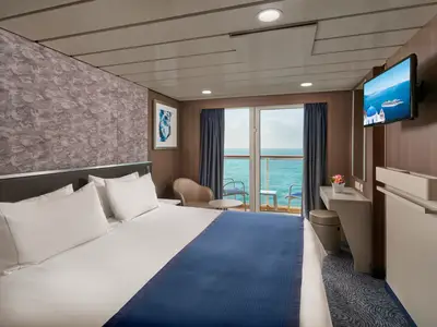 Helle Balkonkabine der Norwegian Spirit. Grosses Bett mit weisser Bettwäsche und blauem Läufer. Links Glastür zum Balkon mit Meerblick. Rechts Schreibtisch, Sessel, Wandfernseher.