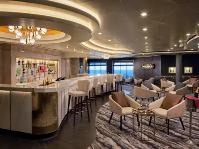 Elegante Magnum's Champagne Bar an Bord der Norwegian Spirit. Eine lange Bar, Barhocker und gemütliche Sitzgruppen dominieren den Raum. Im Hintergrund ist Meerblick durch Fenster zu sehen. Eine Champagnerflasche steht bereit.