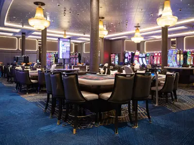 Ein elegantes Casino an Bord der Norwegian Spirit, mit beleuchteten Spielautomaten im Hintergrund und mehreren Roulette-Tischen im Vordergrund, umgeben von dunklen Barhockern. Große goldene Kronleuchter hängen von der Decke, die mit vielen kleinen Lichtern versehen ist.