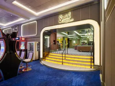 Das beleuchtete Eingangsportal zum Social Comedy & Night Club an Bord der Norwegian Spirit mit Treppe und goldenem Schriftzug. Links sind Reihen von Spielautomaten zu sehen, rechts moderne Wandpaneele.
