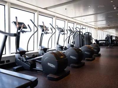 Geräumiger Fitnessbereich an Bord der Norwegian Spirit mit einer Reihe moderner Crosstrainer und Hanteln. Große Fensterfronten bieten Tageslicht und einen Blick nach draußen. Der Boden ist mit dunklem Sportbodenbelag ausgelegt.