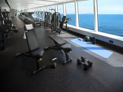 Blick auf den modernen Fitnessraum an Bord der Norwegian Spirit mit grossen Fenstern zum Meer. Verschiedene Hanteln, Hantelbänke und Kardiogeräte sind sichtbar, bereit für das Training mit atemberaubender Aussicht.