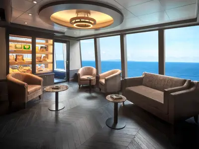 Eine elegante Lounge mit Ledersesseln und einem Sofa sowie einem Humidor mit Zigarrenkisten an Bord der Norwegian Spirit. Große Fenster bieten einen weiten Blick auf das blaue Meer und den Himmel.
