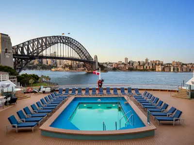Blick über den Poolbereich auf dem Sonnendeck der Norwegian Spirit: blaue Liegestühle, leerer Pool. Im Hintergrund: Sydney Harbour Bridge und Stadt-Skyline.