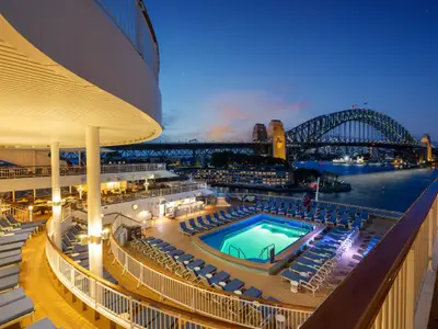 Das beleuchtete Pooldeck der Norwegian Spirit bei Abenddämmerung. Zahlreiche Liegestühle und ein Pool sind zu sehen, mit der Sydney Harbour Bridge und der Skyline im Hintergrund.