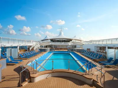 Das sonnige Pooldeck der Norwegian Spirit mit großem Schwimmbecken. Blaue Liegestühle säumen das Holzdeck. Im Hintergrund ist eine überdachte Bar unter blauem Himmel zu sehen.