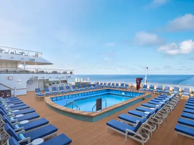 Eine große Poolanlage mit vielen blauen Liegestühlen auf dem Sonnendeck der Norwegian Spirit, dahinter das weite Meer unter blauem Himmel. Eine Bar ist im Hintergrund sichtbar.