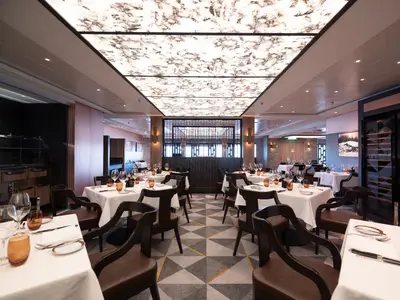 Innenansicht des eleganten Spezialitätenrestaurants Cagney's an Bord der Norwegian Spirit. Weiß gedeckte Tische, dunkle Holzstühle. Eine markante Lichtdecke mit Marmoroptik und eine Weinwand prägen den modernen Raum.