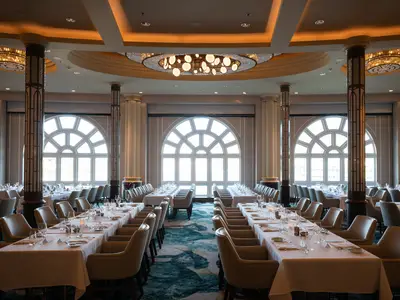 Eleganter Hauptspeisesaal an Bord der Norwegian Spirit mit festlich gedeckten langen Tischen und bequemen Stühlen. Helle Bogenfenster und dekorative Säulen prägen den offenen Raum. Stilvolle Kronleuchter spenden warmes Licht.