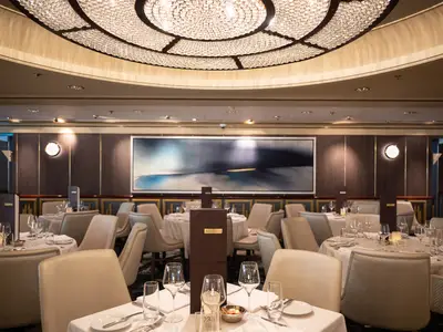 Das elegante Hauptrestaurant Windows an Bord der Norwegian Spirit mit vorbereiteten Tischen, Gläsern und Kerzen. Ein funkelnder Deckenleuchter und ein abstraktes Wandgemälde prägen das luxuriöse Ambiente.