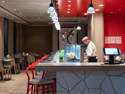 Ein Koch bereitet Sushi an der Theke eines Spezialitätenrestaurants der Norwegian Spirit. Frische Zutaten in Vitrine. Im Hintergrund warten gedeckte Tische und Stühle auf Gäste.
